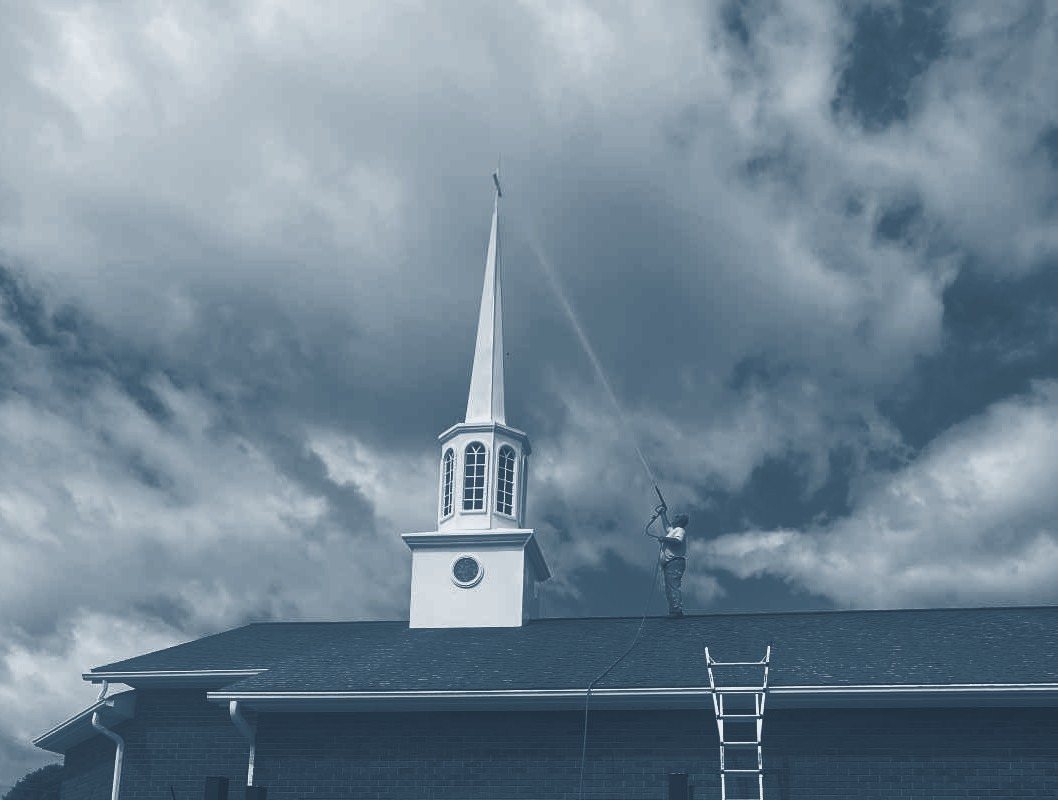 A crew member on a church roof washing beside the steeple, ladder set against the eave