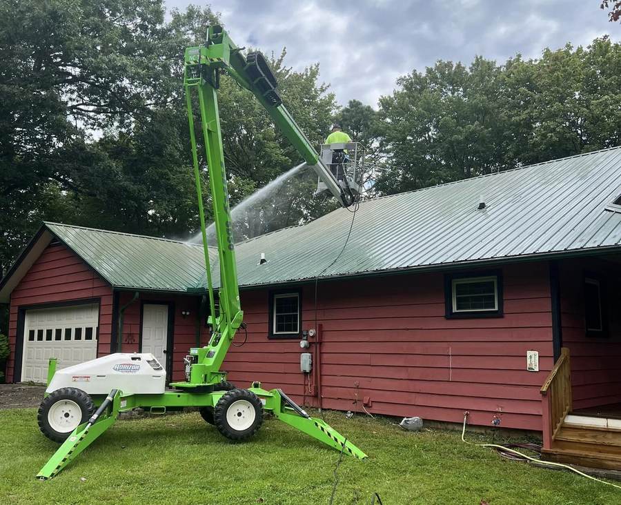 A tracked boom lift with the basket raised over the metal roof of a red barn style building